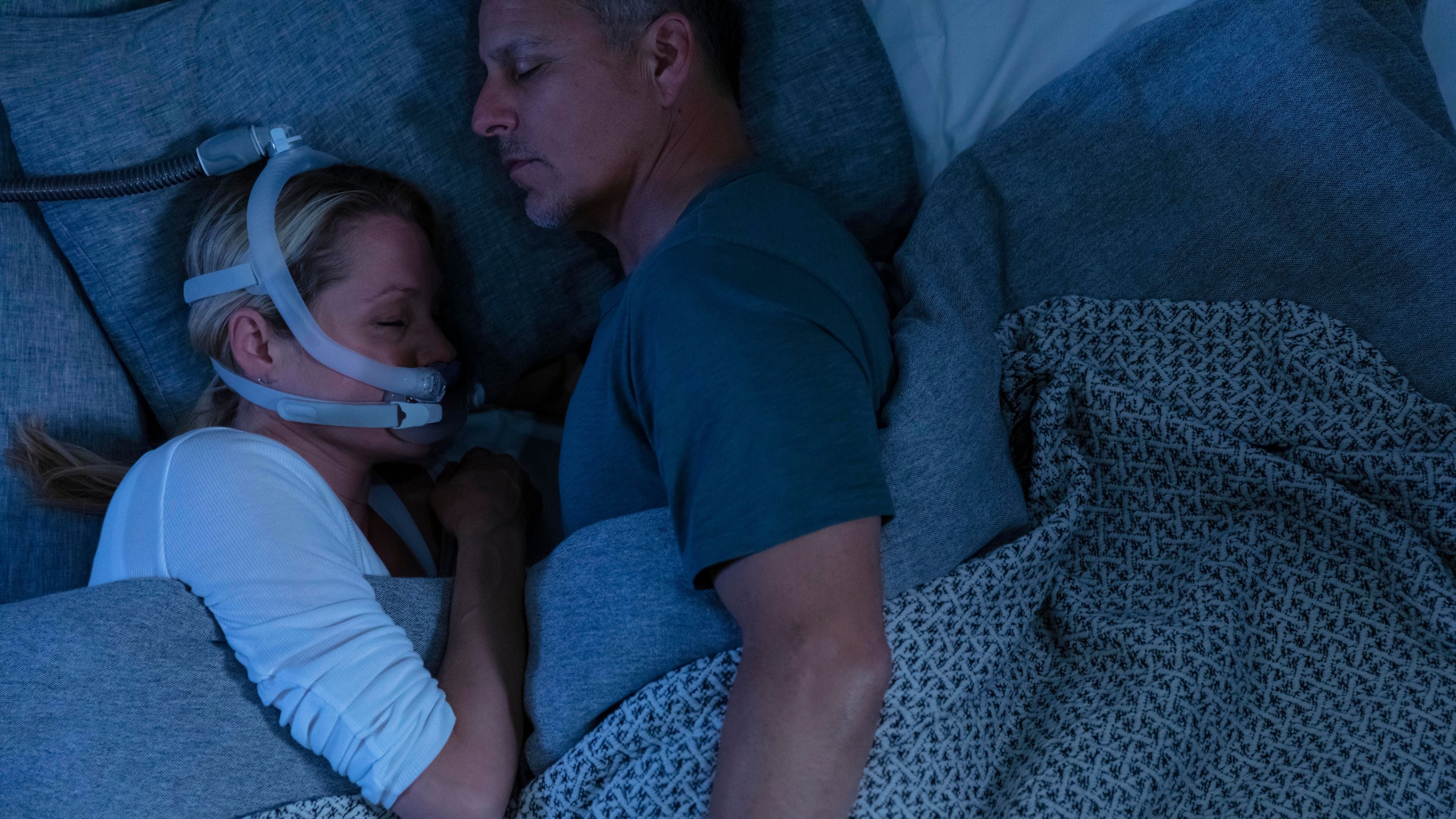 Woman wearing an X30i CPAP mask lying next to a male partner in bed covered in dark coloured sheets.