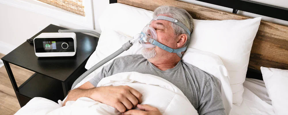 Man wearing a full face mask lying in bed connected to a  iBreeze CPAP machine  device on a nightstand.