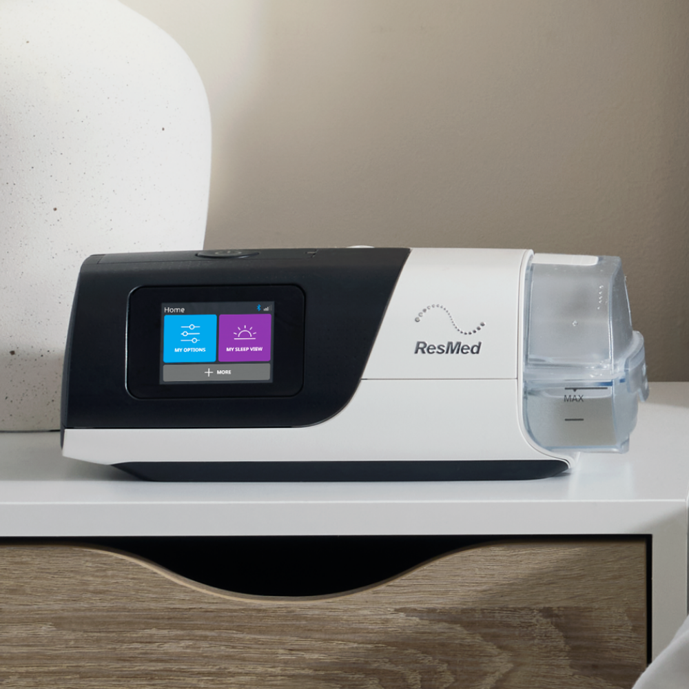 Resmed AirSense 11 CPAP Machine  sitting on a white and wood counter with a screen displaying icons on neutral backdround