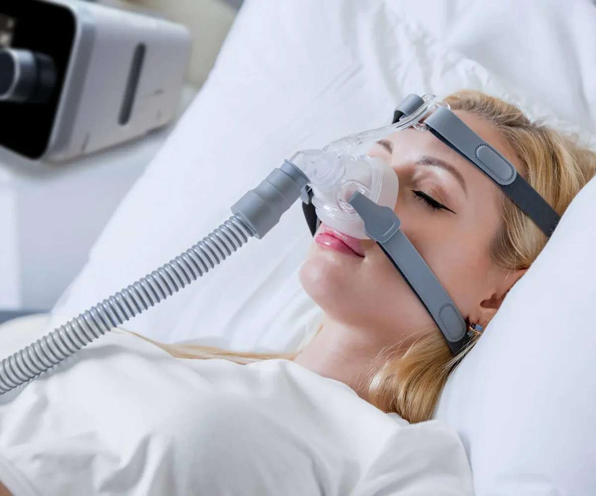 A woman sleeps comfortably in her bedroom wearing a CPAP mask connected by tubing, with the iBreeze machine resting on the bedside table.