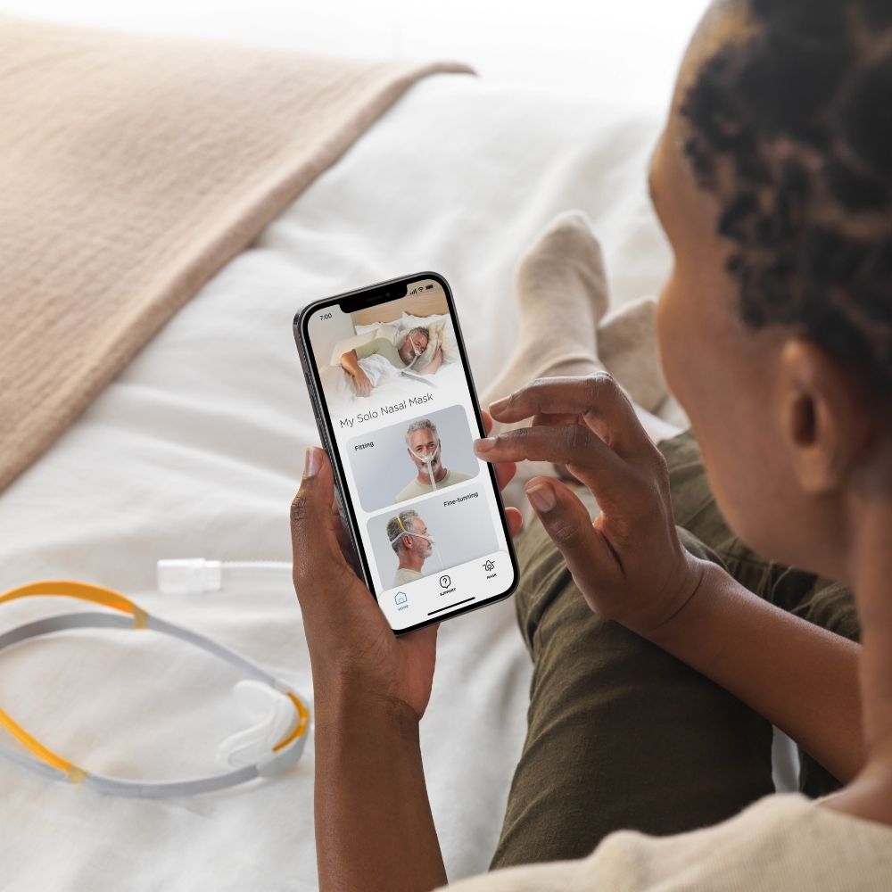 Person using a mobile phone to access the Fisher & Paykel MyMask app on a bed with the Solo CPAP mask nearby