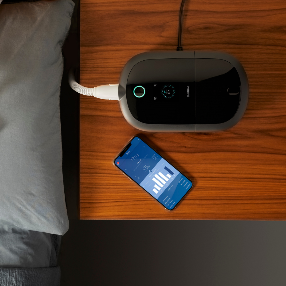 Philips DreamStation 2 CPAP machine on nightstand beside a smartphone displaying the DreamMapper app interface