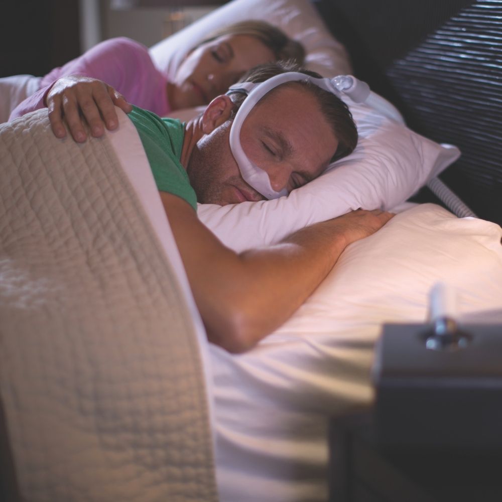Two people sleeping in bed, one person using using the Philips DreamWear Nasal Mask while sleeping on their stomach