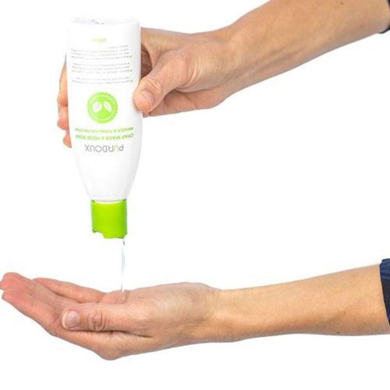 Hand holding a white and green bottle of Purdoux soap with liquid soap being dispensed onto another hand