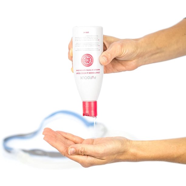 Hand holding a white and red bottle of Purdoux soap with liquid soap being dispensed onto another hand