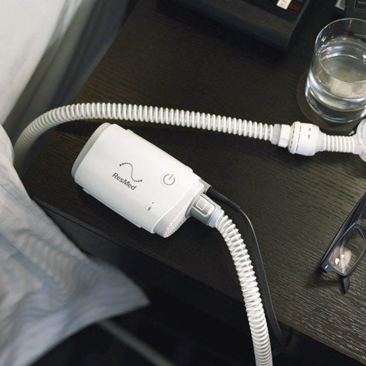 Resmed AirMini CPAP machine with hose on a nightstand next to a glass of water and glasses
