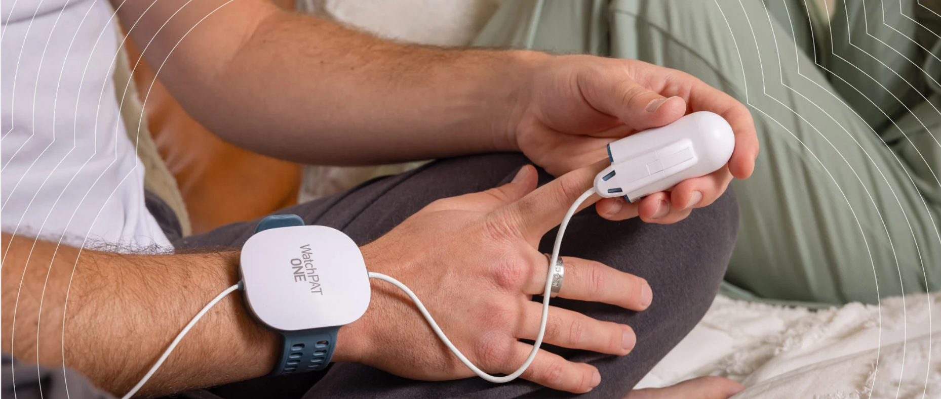 A person sitting cross legged indoors in a bedroom setting wearing the watchpat one home sleep test on wrist and holding the small white fingertip over index finger