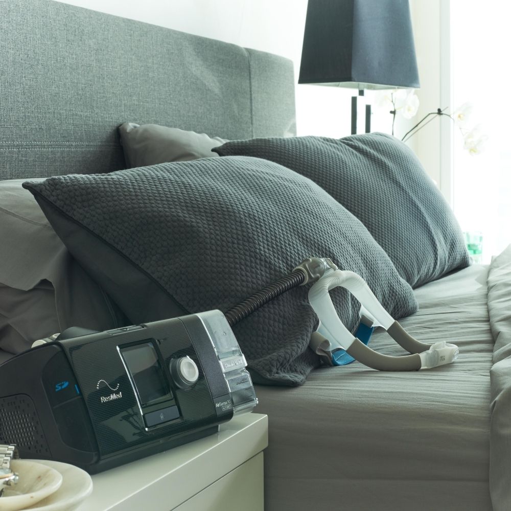 CPAP machine on a nightstand next to a bed with pillows and a mask