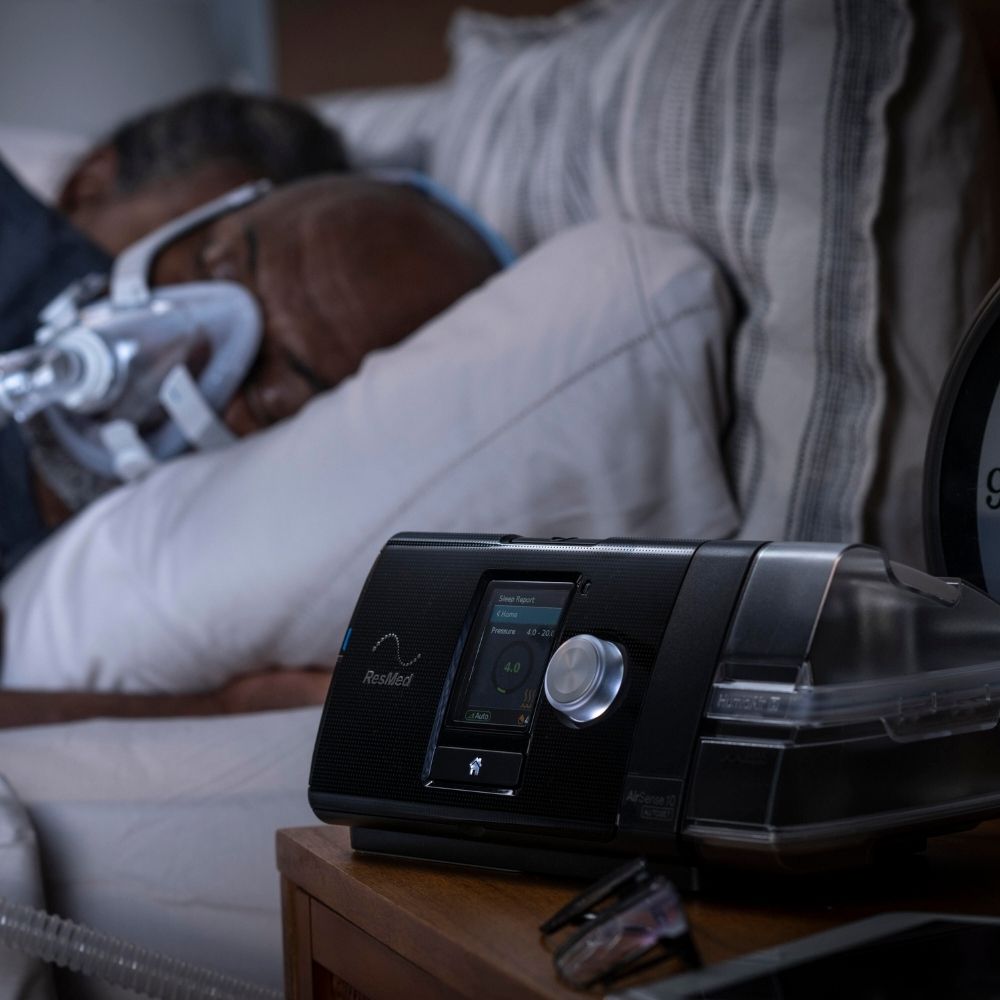 AirSense 10 CPAP machine on nightstand with person sleeping beside it with a full face mask on