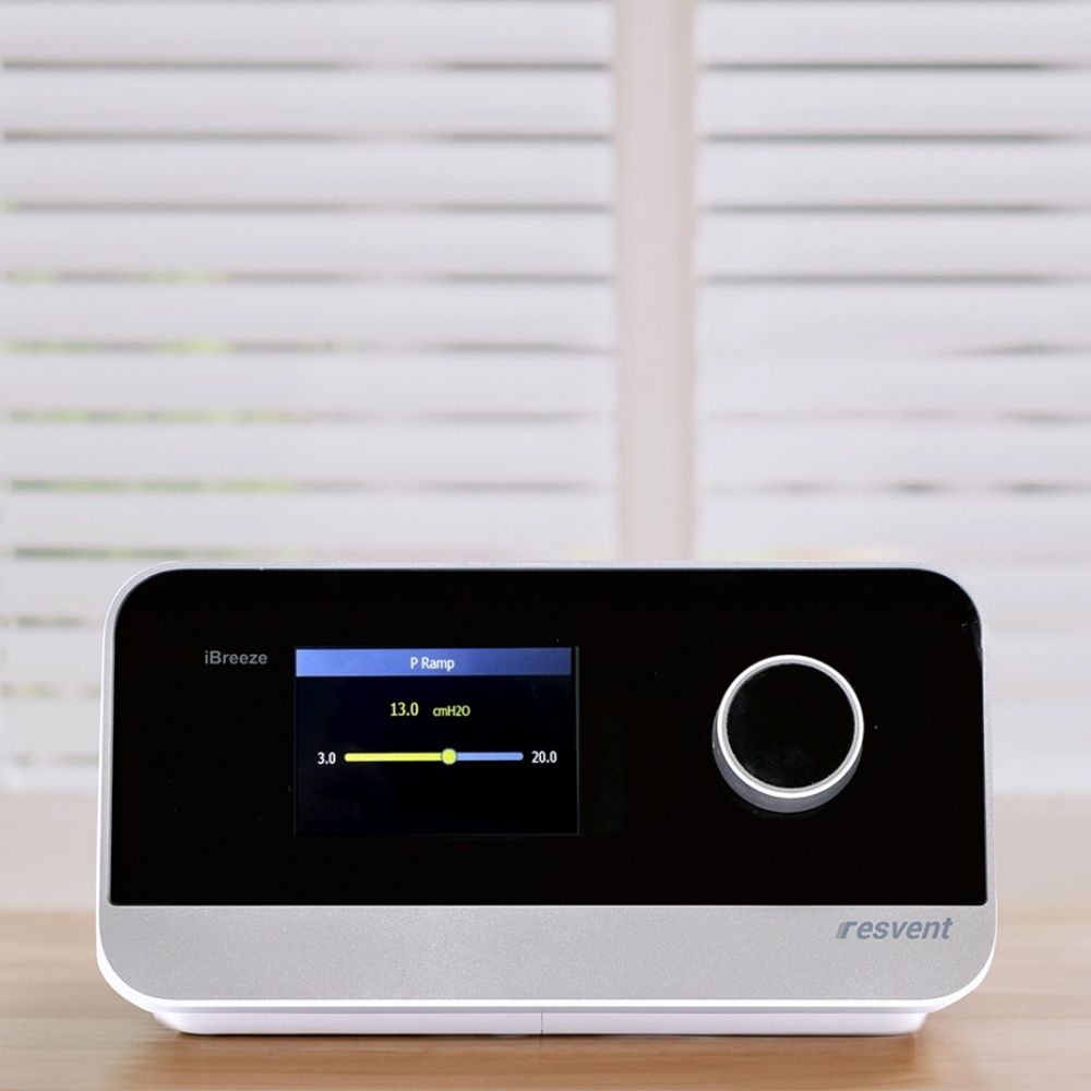 Resvent iBreeze CPAP machine on a wooden surface with white blinds in the background