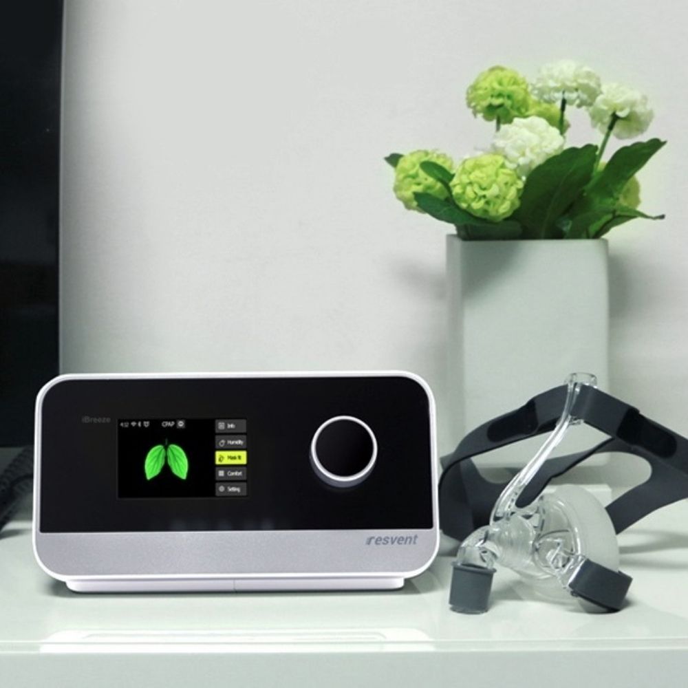 Resvent iBreeze CPAP machine and mask on a white table with flowers in a vase behind it